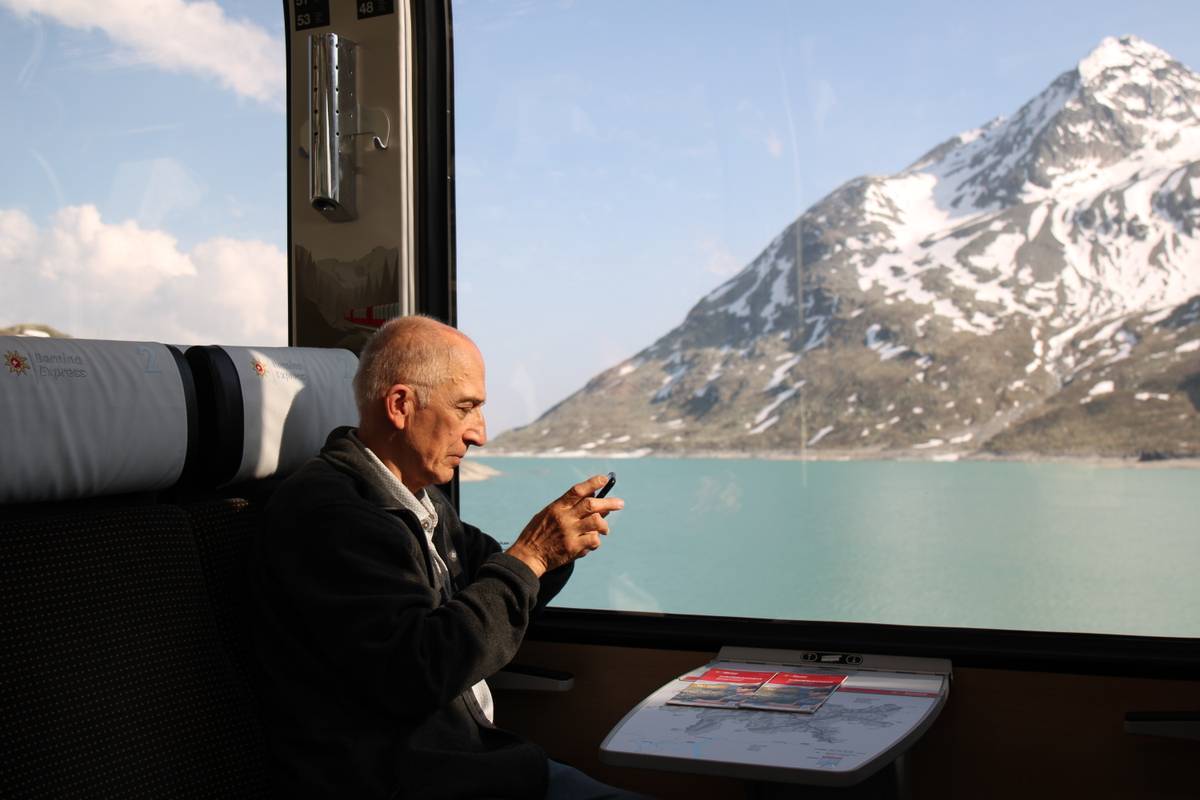 My dad sits in the train checking his phone. Outside the tall window is a giant lake and a large mountain.