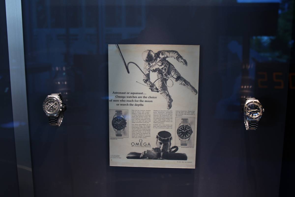 Two watches are pictured in a display case with a magazine ad which features them both placed between them